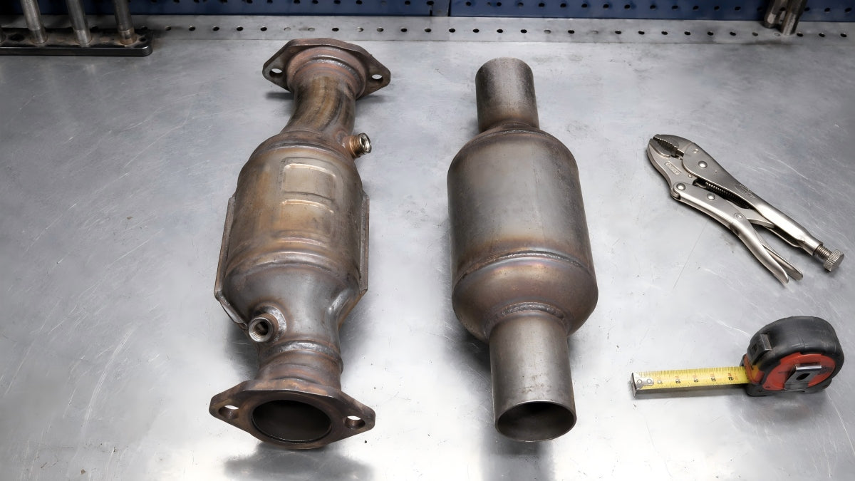 Direct-fit catalytic converter with flanges and O2 sensor bungs next to universal catalytic converter on metal workbench, showing size difference and installation complexity comparison